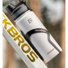 A white insulated stainless steel thermal water bottle with a black cap is shown next to a yellow and black branded item.