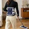 A woman wearing a black long-sleeved top and beige pants demonstrates an adjustable lumbar support back brace.
