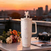 A white insulated thermos mug with a pink floral pattern sits on a table against a sunset cityscape.