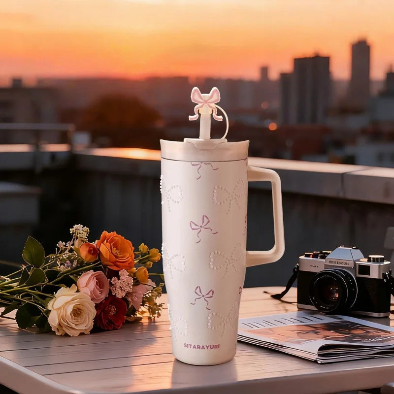 A white insulated thermos mug with a pink floral pattern sits on a table against a sunset cityscape.