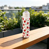 A white tumbler decorated with red strawberries and green leaves sits on a wooden table outdoors.