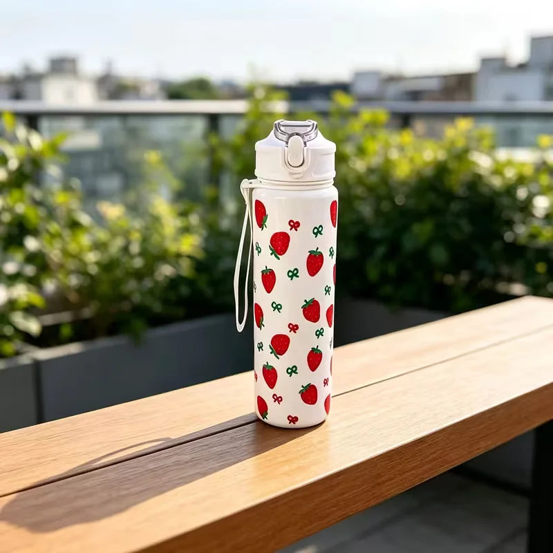 A white tumbler decorated with red strawberries and green leaves sits on a wooden table outdoors.
