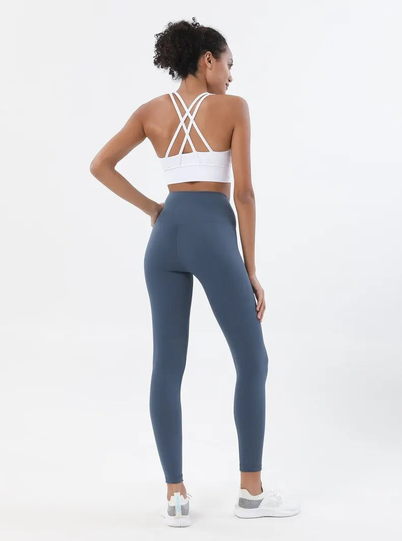 Eine Frau in einem weißen Sport-BH und dunkelblauen Yoga-Hosen posiert vor einem hellen Hintergrund.