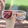 A hand holds a pair of pink photochromic square frame sunglasses with UV400 protection against a blurred green outdoor backgr