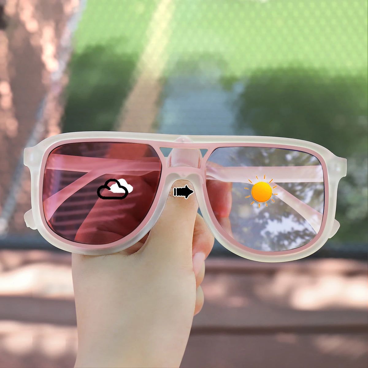 A hand holds a pair of pink photochromic square frame sunglasses with UV400 protection against a blurred green outdoor backgr