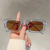 Une main tient des lunettes de soleil rectangulaires rétro en plastique transparent avec des verres ambrés.