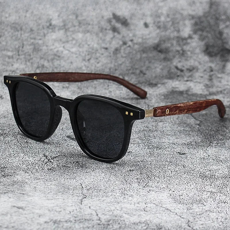 Black sunglasses with dark lenses and a brown wood grain frame resting on a gray textured surface.