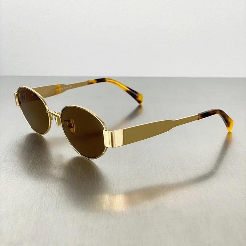 Gold-toned metal oval sunglasses with brown tinted lenses resting on a gray surface.