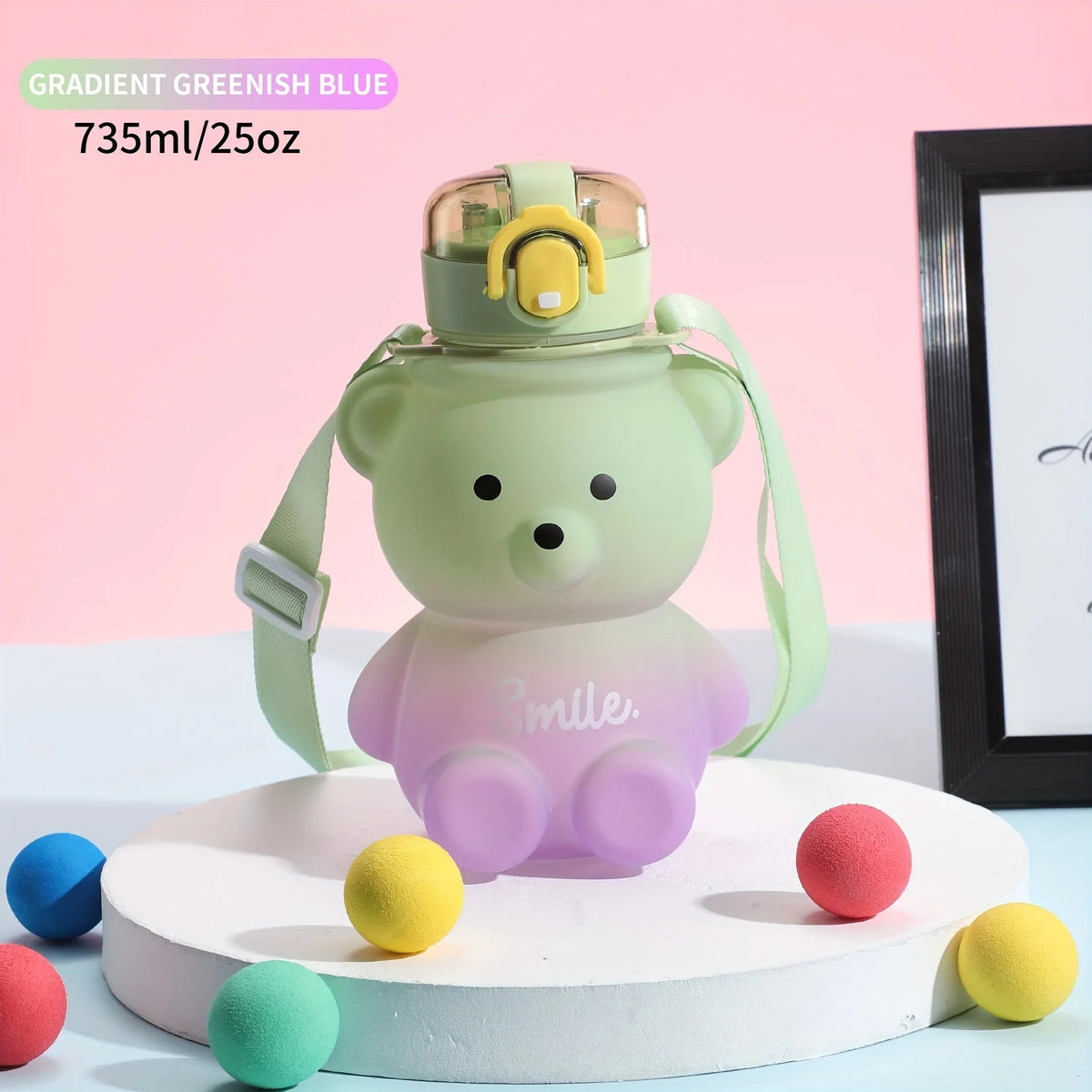A light green bear-shaped water bottle with a straw and strap is displayed on a white stand surrounded by colored balls.