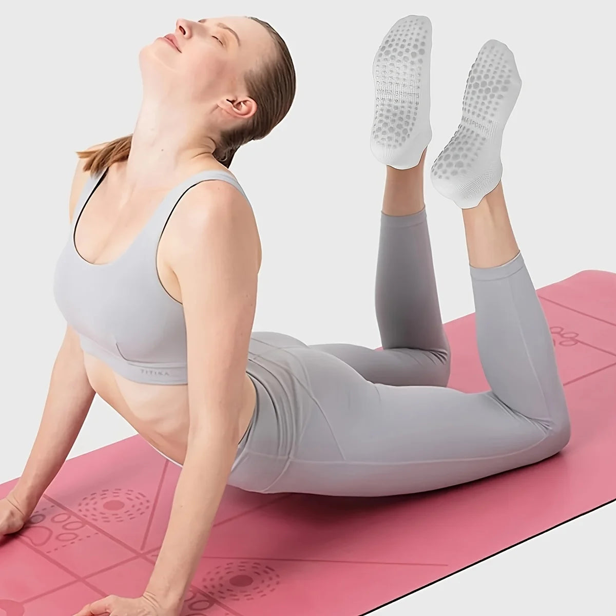 A woman wearing gray athletic wear is performing a stretch on a pink mat, demonstrating Grip Pilates Socks for Yoga and Barre