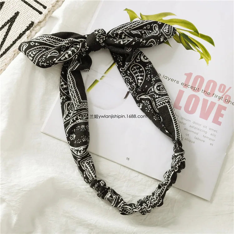 Tête en tissu paisley noir bohème noué en nœud, posée sur une surface blanche avec des feuilles vertes.