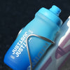A blue cycling water bottle with a metal clip is shown resting on a pink and white bicycle frame.