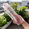 A hand holds a clear reusable water bottle with a pink and green strawberry pattern outdoors.