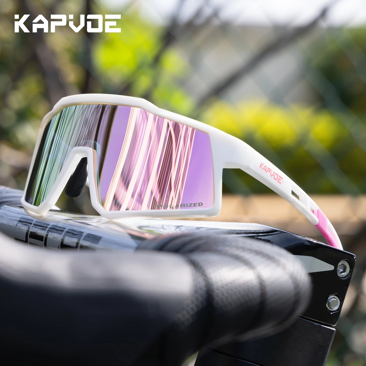 A pair of white Kapvode polarized sports sunglasses with pink mirrored lenses resting on a black grip.