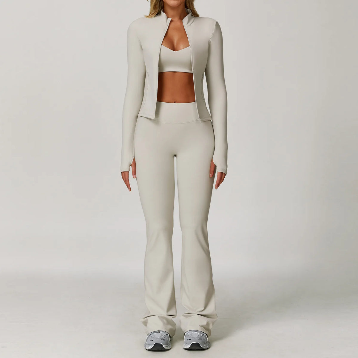 A woman models a beige 3-piece yoga sports set consisting of a cropped top, long-sleeved jacket, and matching pants.