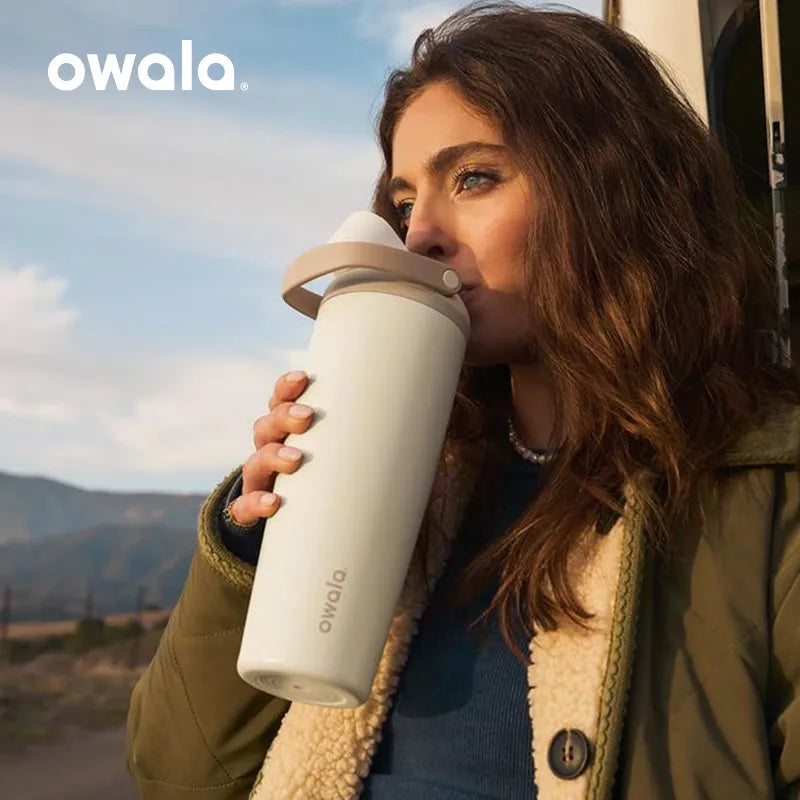 A woman holds an Owala insulated stainless steel travel mug outdoors against a scenic, hazy landscape.