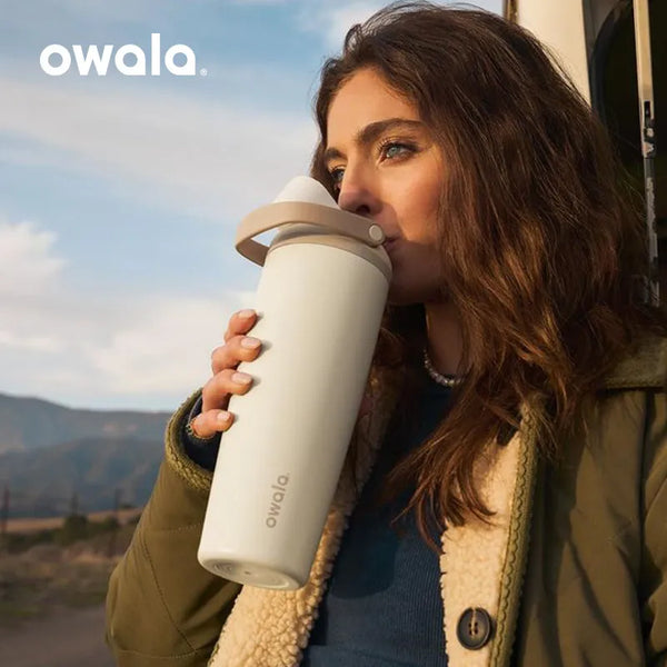 A woman holds an Owala insulated stainless steel travel mug outdoors against a scenic, hazy landscape.