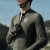 A male cyclist wearing a dark olive performance jersey and black gloves, looking forward while riding a bicycle.