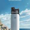 A white insulated sports bottle with a black cap is displayed outdoors on a rocky surface with white flowers and blue water i