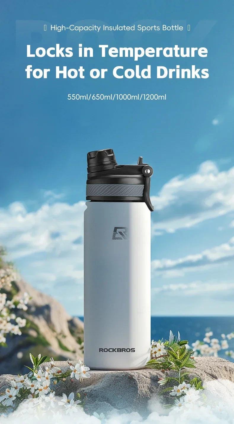A white insulated sports bottle with a black cap is displayed outdoors on a rocky surface with white flowers and blue water i