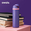 Lila Owala isolierte Wasserflasche mit rosa Deckel auf rosa Oberfläche neben gestapelten Papieren.