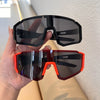 A hand holds a pair of black and orange cycling sunglasses against a white background.