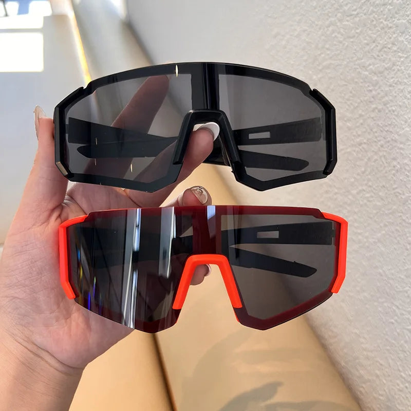 A hand holds a pair of black and orange cycling sunglasses against a white background.
