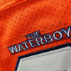 Nahaufnahme eines orangefarbenen Fußballtrikots mit blauem gesticktem Text, der 'THE WATERBOY' lautet