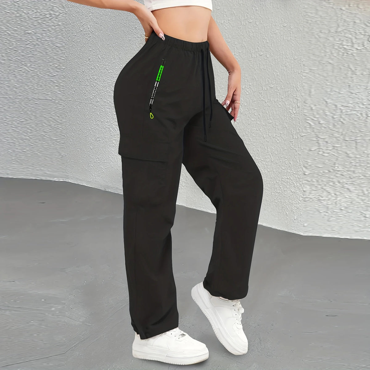 A woman wearing black drawstring cargo pants and a white top standing against a gray background.