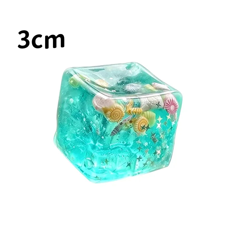 A translucent teal cube filled with glitter and small decorative elements, used as a squishy stress relief fidget cube.