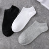 A stack of three ankle socks in black, white, and gray are displayed on a gray textured surface.
