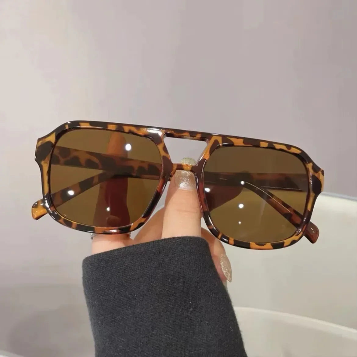 Una mano sostiene unas gafas de sol cuadradas con patrón tortuga y lentes marrones contra un fondo claro.
