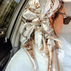 A close-up of a woman wearing an elegant satin square scarf tied around her hair, featuring brown and beige floral patterns.
