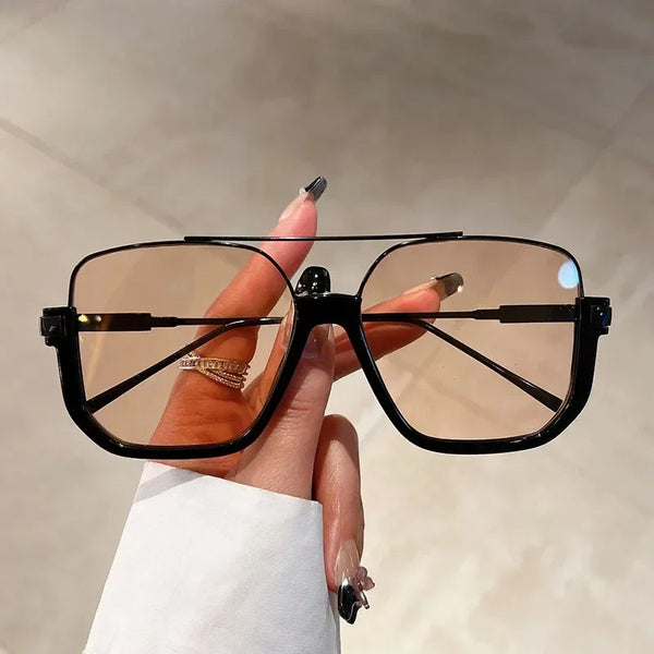 A hand holds a pair of black vintage oversized square sunglasses with brown tinted lenses against a light background.