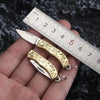 A brass folding knife keychain is shown resting on a ruler against a dark background.