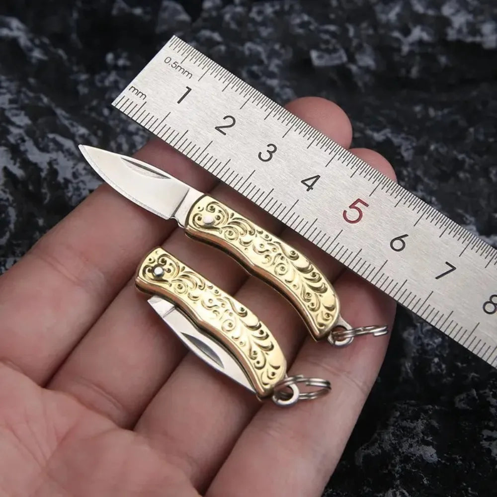 A brass folding knife keychain is shown resting on a ruler against a dark background.