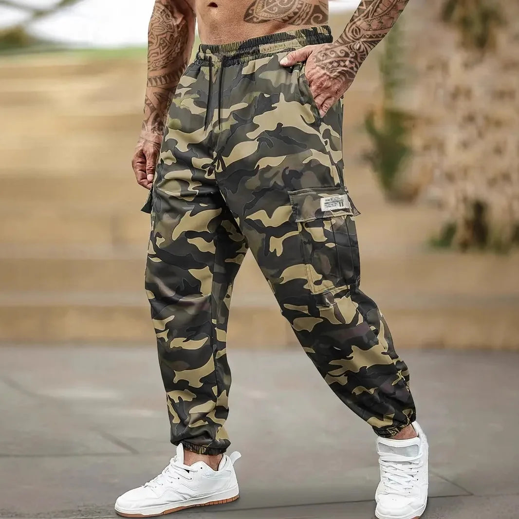 Un homme portant un pantalon cargo camouflage et un débardeur, debout à l'extérieur.