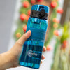 A hand holds a blue glass water bottle with a black cap and tea infuser against a blurred green Christmas wreath background.