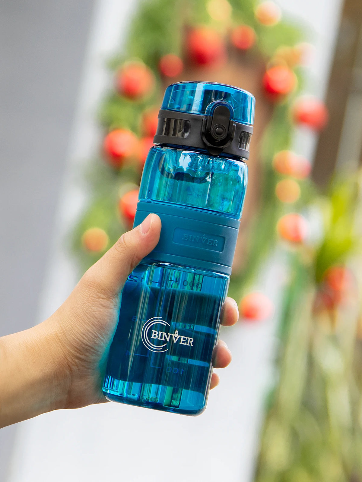 A hand holds a blue glass water bottle with a black cap and tea infuser against a blurred green Christmas wreath background.