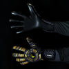 A hand wearing black and gold high-performance goalkeeper gloves is extended against a dark background.