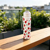 A white reusable water bottle decorated with a pattern of red strawberries and green bows, sitting on a wooden table.