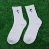 A pair of white knit mid-calf sports socks with black crosses on the toes lying on green grass.