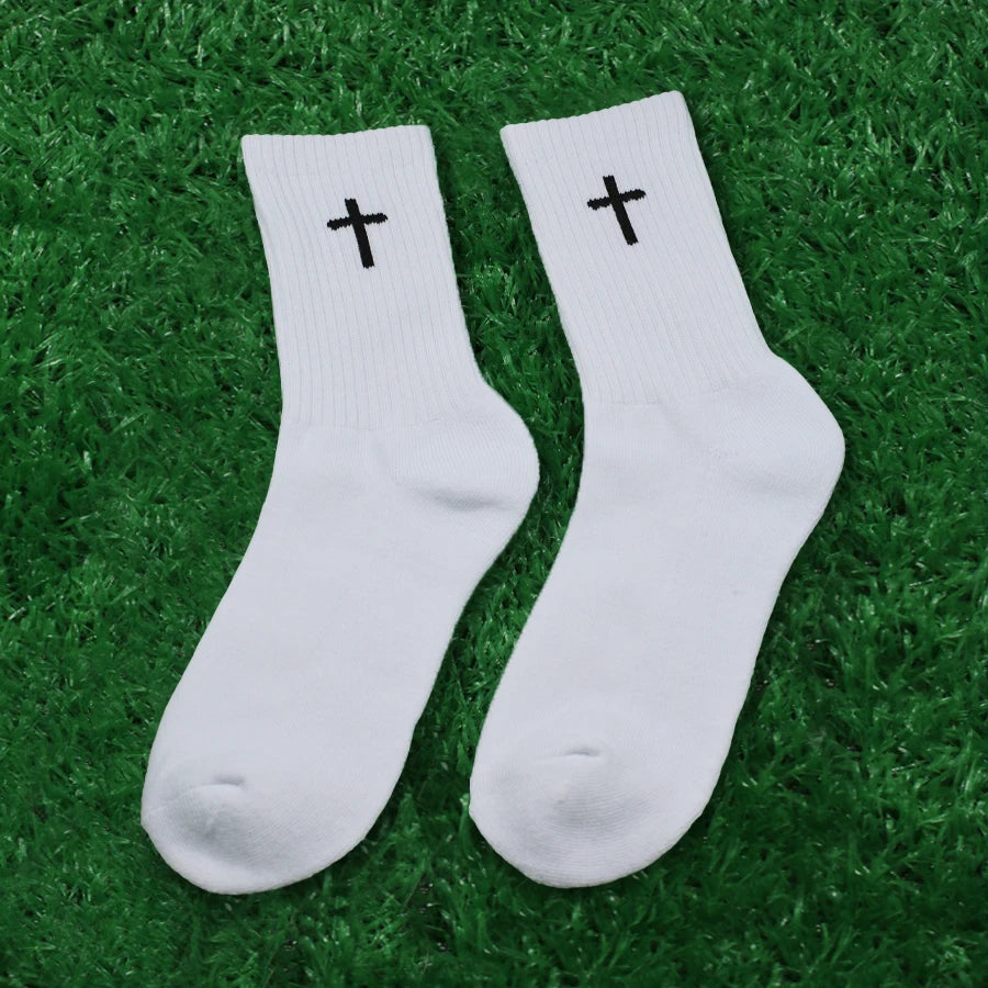 A pair of white knit mid-calf sports socks with black crosses on the toes lying on green grass.