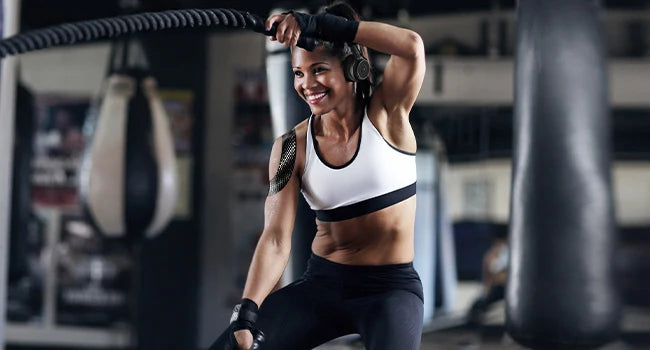 A smiling woman performing a physical exercise with kinesiology tape on her arm in a gym setting.