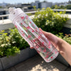 A hand holds a clear reusable water bottle with pink strawberry bow designs outdoors.