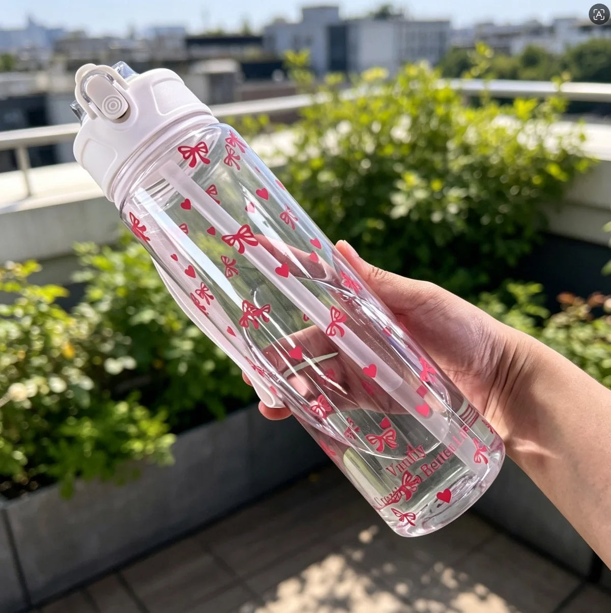 A hand holds a clear reusable water bottle with pink strawberry bow designs outdoors.