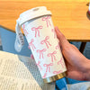 A hand holds a white travel mug with pink ribbon pattern on a wooden table.