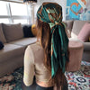 A woman with long brown hair wearing a dark green satin square scarf tied around her head in a relaxed indoor setting.