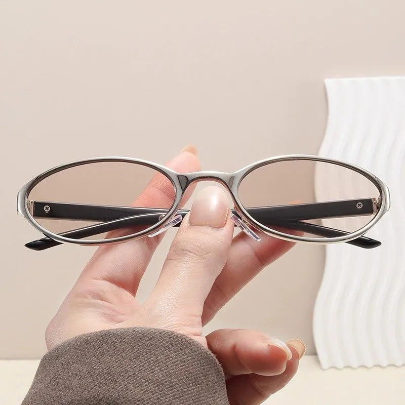 A hand holds a pair of retro oval sunglasses with a silver metal frame against a light background.
