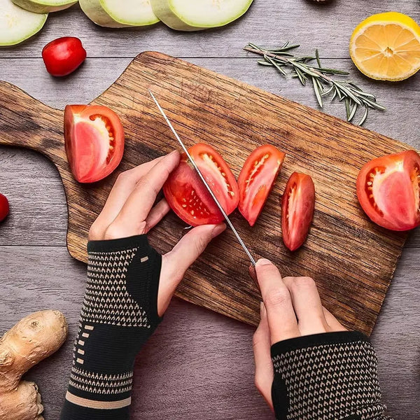 Hands wearing black knitted wrist supports are slicing fresh tomatoes on a wooden cutting board.